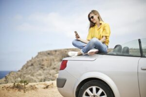 Jeune femme souriante assise sur une voiture sur la plage de Mgarr, Malte, utilisant un smartphone.